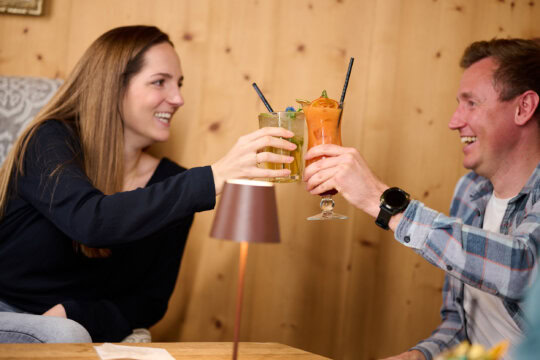 Zwei Personen stoßen mit Getränken in einer gemütlichen Umgebung an.
