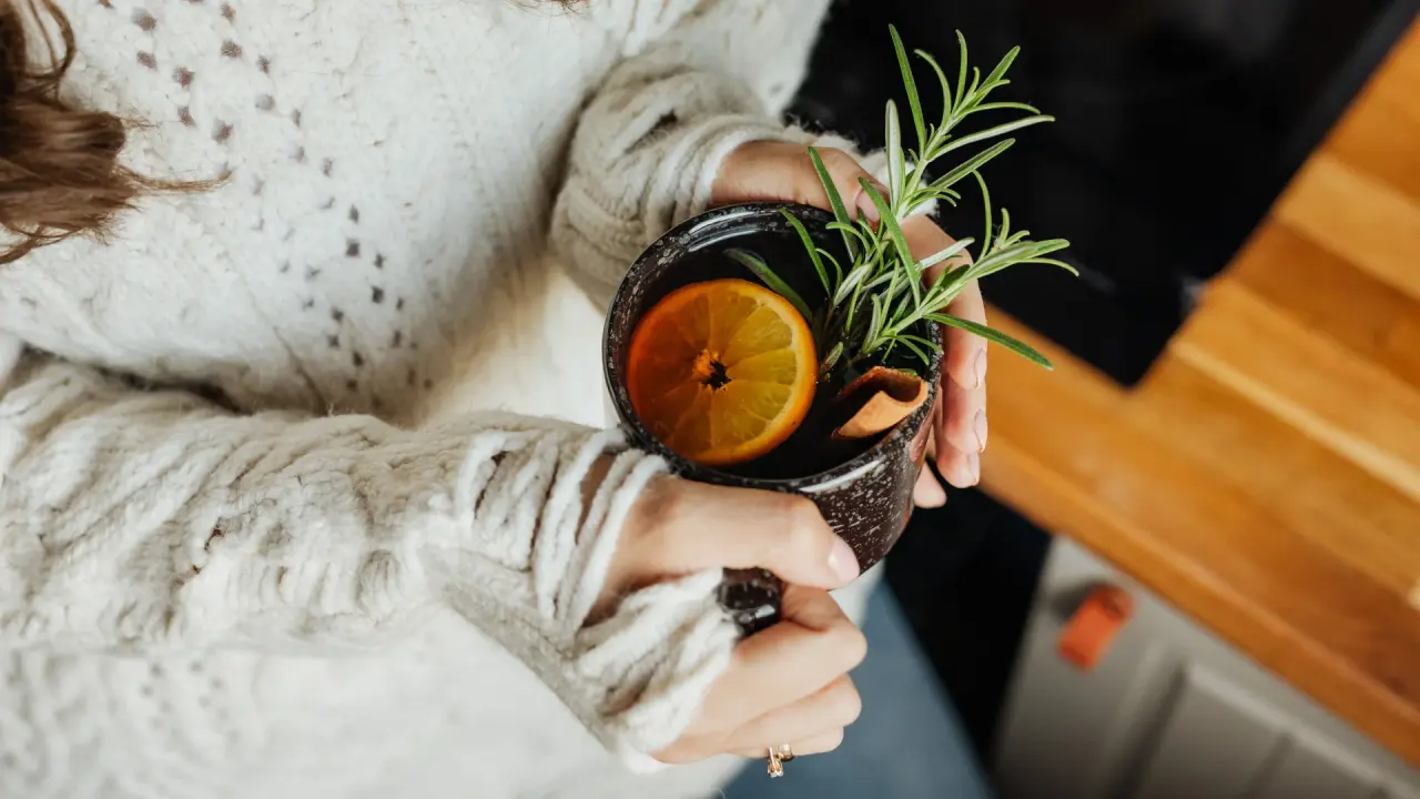 Person hält Tasse mit Heißgetränk, Orange und Rosmarinzweig.